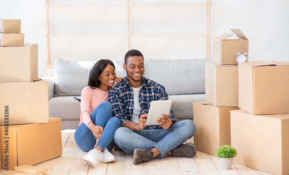 Cheerful black couple with touch pad to search for household goods for their new home online, using tablet on moving day Cheerful black couple with touch pad to search for household goods for their new home online, using tablet on moving day