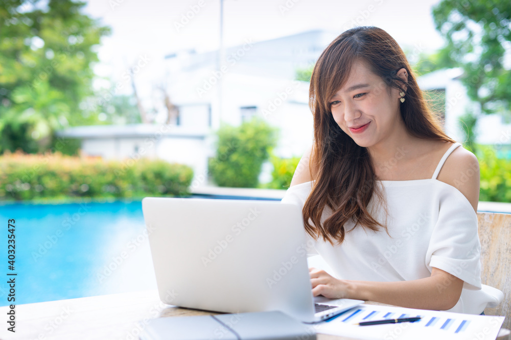 An Asian female office worker taking video calls, online conferences, smiling and greeting colleagues through her laptop at the poolside table, the concept of a home-made video call via the Internet. An Asian female office worker taking video calls, online conferences, smiling and greeting colleagues through her laptop at the poolside table, the concept of a home-made video call via the Internet.