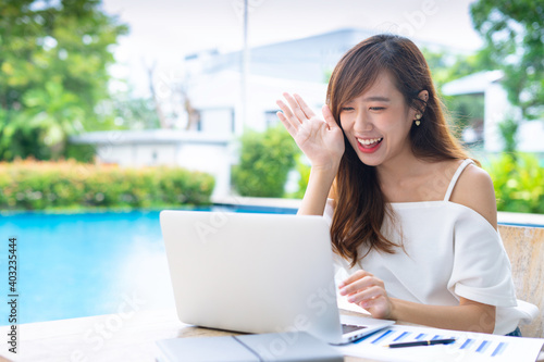 An Asian female office worker taking video calls, online conferences, smiling and greeting colleagues through her laptop at the poolside table, the concept of a home-made video call via the Internet. Free Download An Asian female office worker taking video calls, online conferences, smiling and greeting colleagues through her laptop at the poolside table, the concept of a home-made video call via the Internet. - GFXLoad