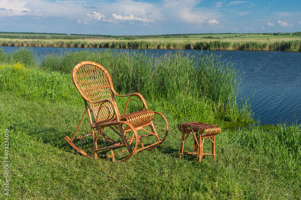 Empty wicker rocking-chair standing next to small wicker hadnmade stool,  on a Sura riverside waiting for any human to relax