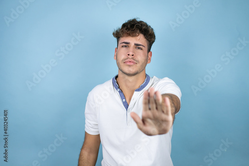 Young handsome man wearing a white t-shirt over blue background serious and doing stop sing with palm of the hand.