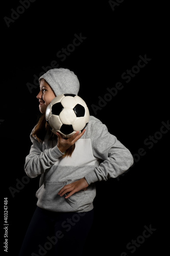 Fototapeta Naklejka Na Ścianę i Meble -  Little girl in gray sweatshirt and two pigtails playing with a soccer ball isolated on black background