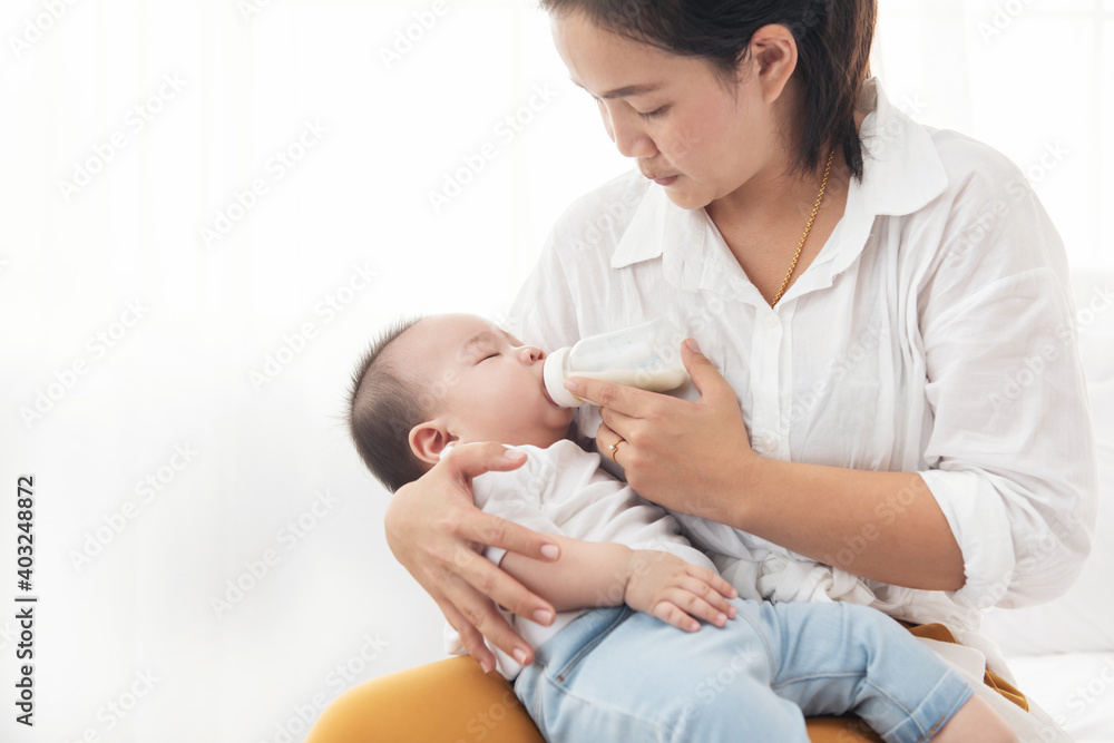 Portrait of a happy asian mother giving bottle feeding to her baby in white bedroom. Baby is drinking milk from a bottle hold by the mother. Portrait of a happy asian mother giving bottle feeding to her baby in white bedroom. Baby is drinking milk from a bottle hold by the mother.