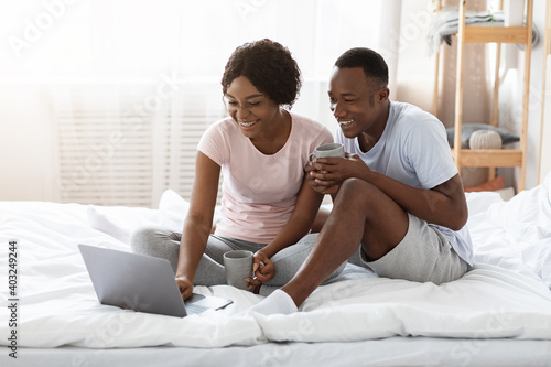 Joyful african couple sitting on bed, watching movie on laptop