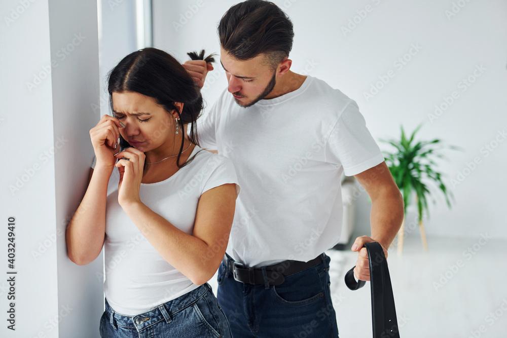 Naklejka premium Violent aggressive young man with belt threatening his girlfriend at home