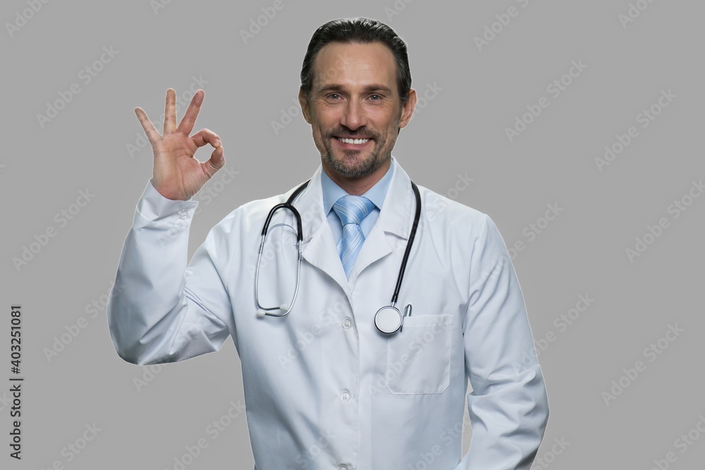 Portrait of handsome doctor showing ok sign. Cheerful male practitioner with stethoscope looking at camera on gray background.