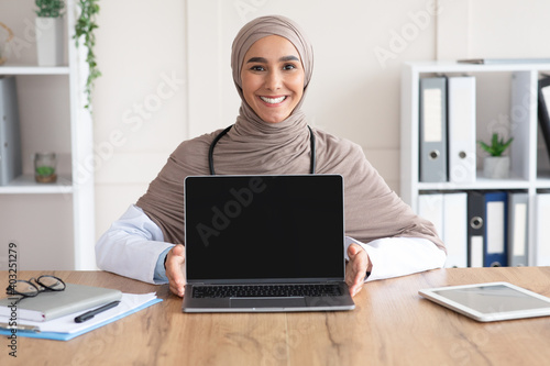 Happy female doctor showing laptop with blank screen, clinic interior
