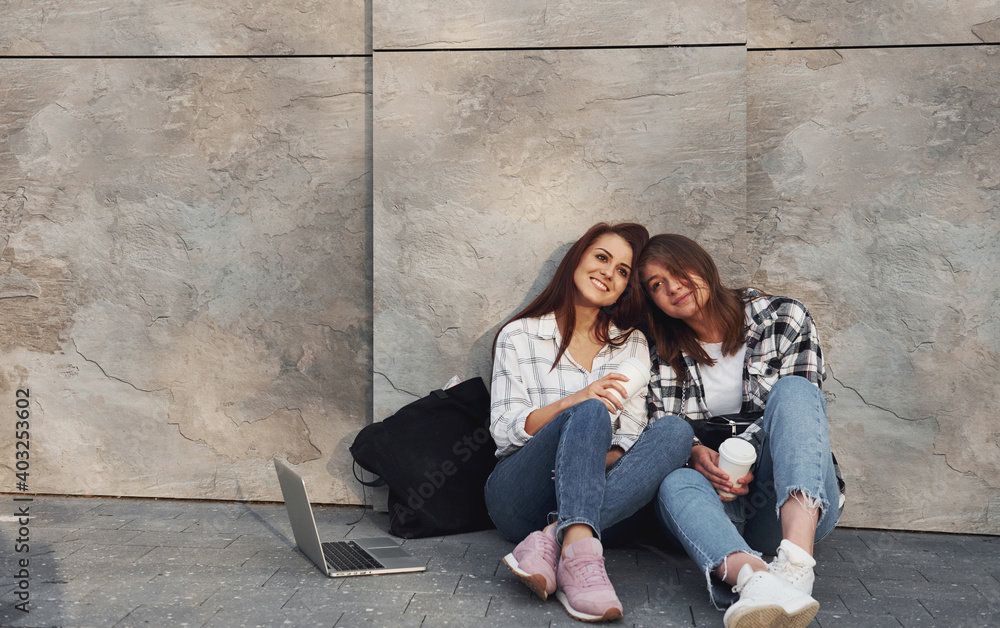 With cup of coffee. Beautiful cheerful friends or lesbian couple together near wall outdoors at daytime