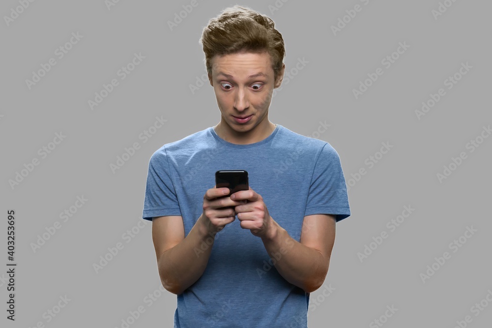 Surprised teenage boy using mobile phone. Trendy teen guy using app on his smartphone against gray background. Surprised teenage boy using mobile phone. Trendy teen guy using app on his smartphone against gray background.