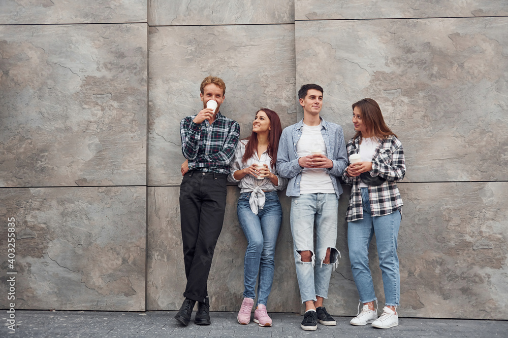 Group of young positive friends in casual clothes standing together against grey wall with cups of drink in hands