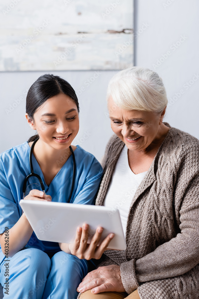 cheerful asian nurse pointing with finger at digital tablet near smiling senior woman cheerful asian nurse pointing with finger at digital tablet near smiling senior woman