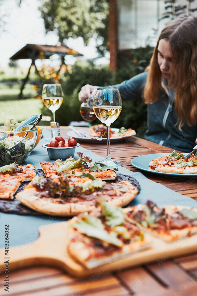 Family and friends having meal - pizza, salads, fruits and drinking white wine during summer picnic outdoor dinner in a home garden. Close up of people sitting at the table in a orchard in a backyard