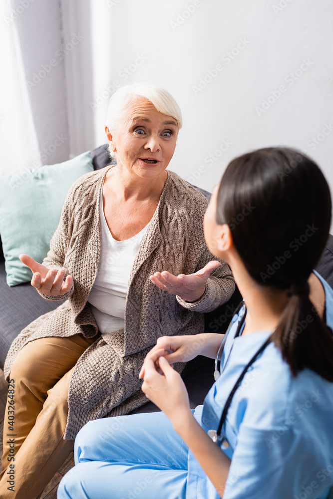 Obraz premium amazed senior woman gesturing during conversation with nurse in nursing home, blurred foreground