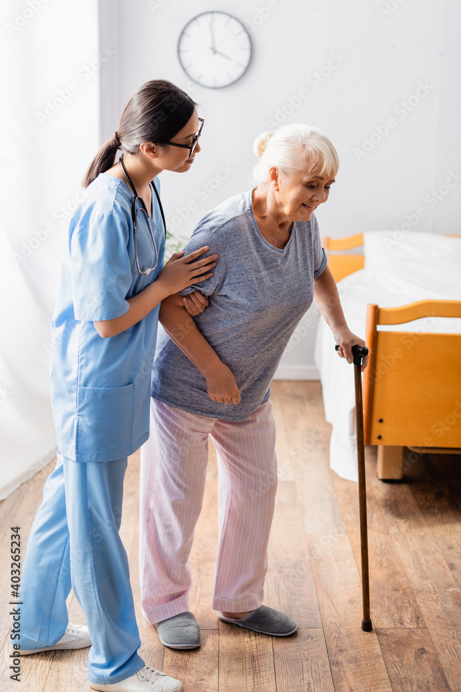 asian nurse helping aged woman walking with stick in hospital