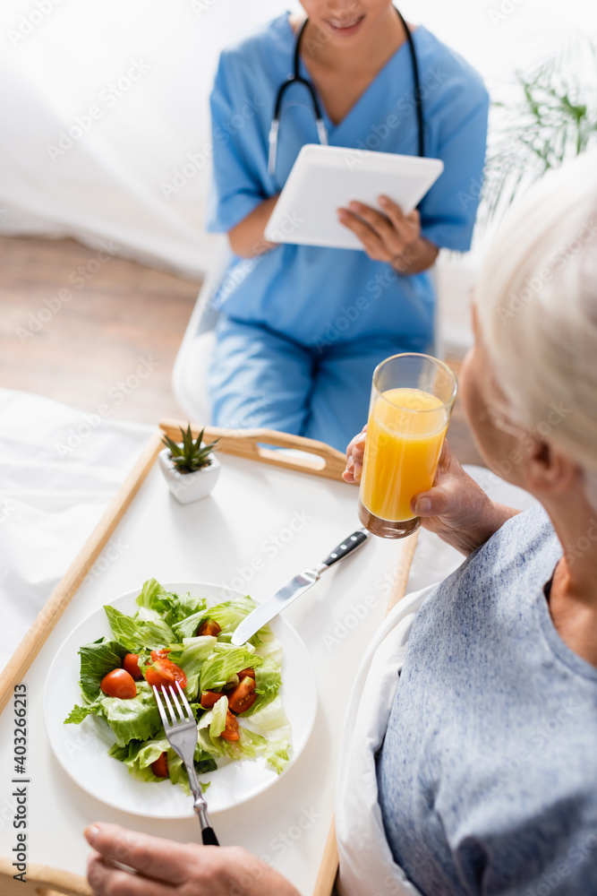 high angle view of senior woman having breakfast near nurse using digital tablet on blurred background high angle view of senior woman having breakfast near nurse using digital tablet on blurred background