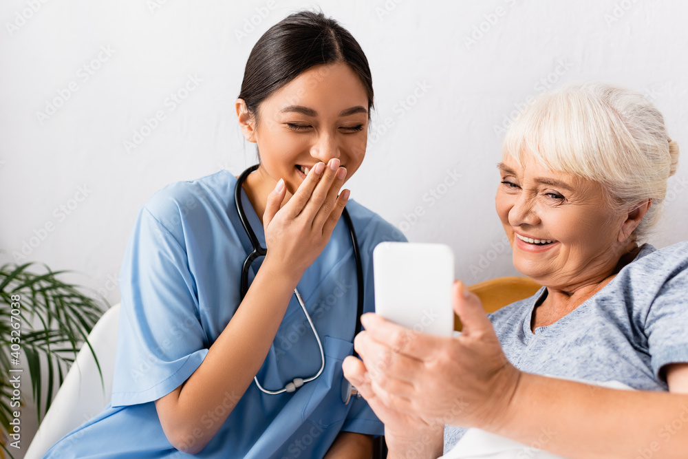 laughing asian nurse covering mouth with hand near cheerful senior woman using mobile phone, blurred foreground laughing asian nurse covering mouth with hand near cheerful senior woman using mobile phone, blurred foreground