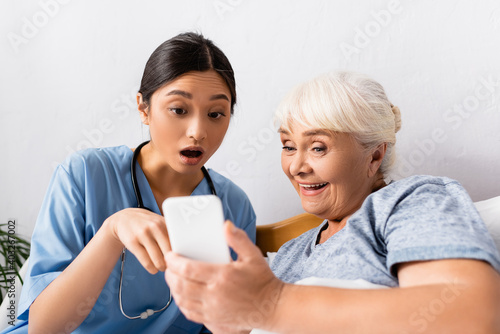 astonished asian nurse pointing at cellphone near excited elderly woman, blurred foreground