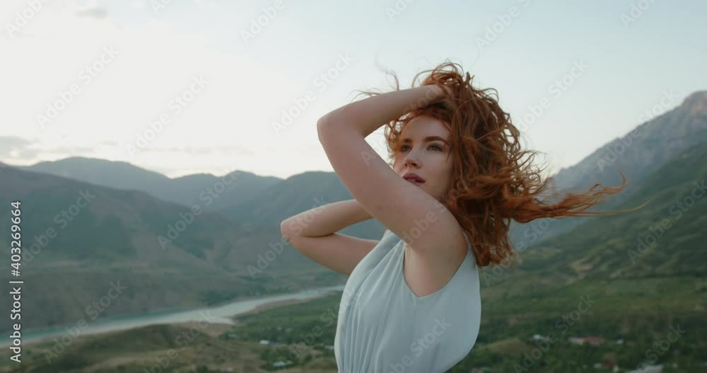 Gorgeous caucasian girl wearing a light dress is posing for a photo shoot on scenic mountain background while wind is blowing her red hair 4k footage Gorgeous caucasian girl wearing a light dress is posing for a photo shoot on scenic mountain background while wind is blowing her red hair 4k footage