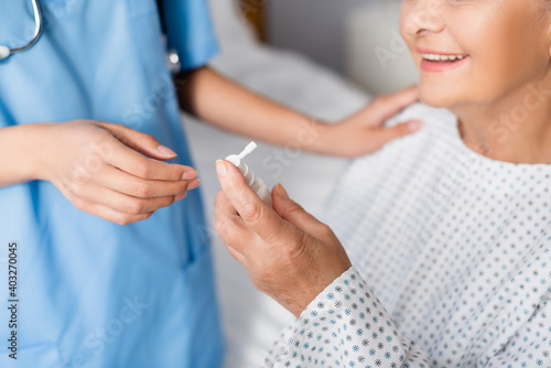 cropped view of aged, smiling woman holding nasal spray near nurse on blurred background