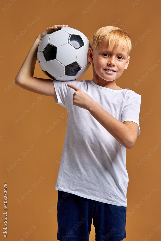 football player child isolated in studio, posing at camera, wearing football uniform, holding ball on shoulders. sport, soccer concept