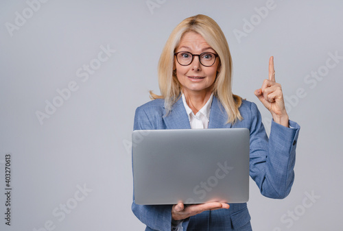 Senior pretty business woman with laptop pointing at copy space isolated over grey background