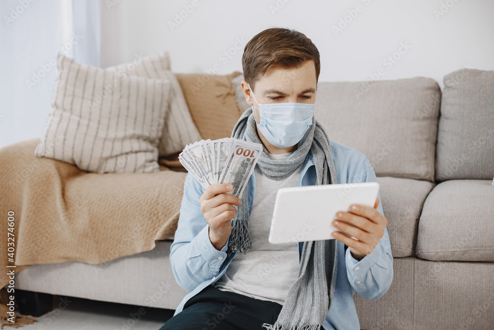 Male in a medical mask. Man sitting in living room at home. Guy have online money on quarantine. Male in a medical mask. Man sitting in living room at home. Guy have online money on quarantine.