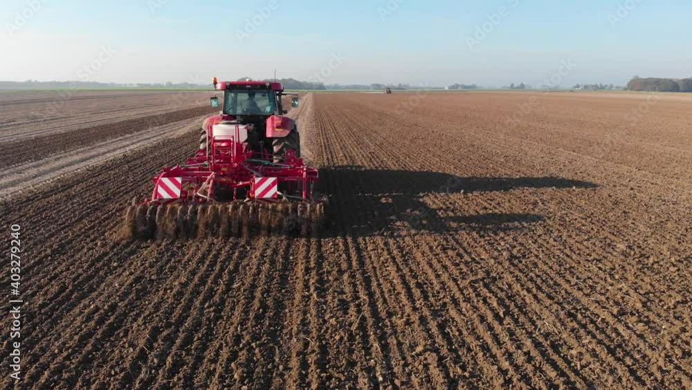 Red tractor plowing brown, earthy, dutch farmland in vertical lines under blue skies Red tractor plowing brown, earthy, dutch farmland in vertical lines under blue skies