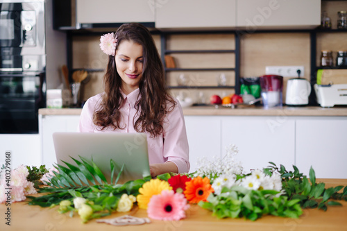 Young brunette woman with long wavy hair working on laptop with flowers on the desk with kitchen in background. Talented florist developing online sales getting ready for workshop.