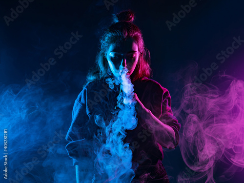 Girl smokes a VAPE on a dark background. Woman lets out clouds of smoke from an electronic cigarette. Girl stands in the middle of the frame and looks at the smoke from the e-cigarette. VAPE shops.