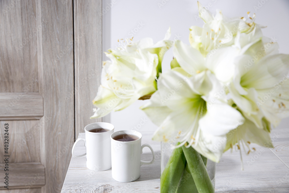 Bouquet of white lilies in a tall glass vase on a beige table against a gray wall.