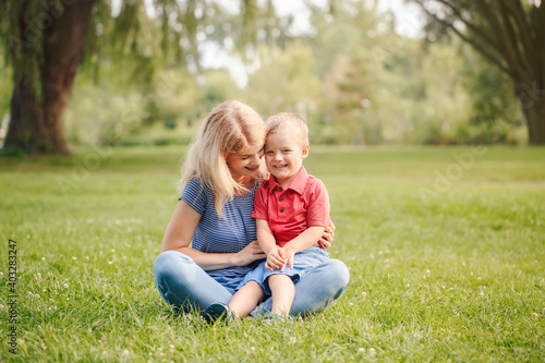 Mothers Day holiday. Young smiling Caucasian mother and laughing boy toddler son sitting on grass in park. Family mom and child hugging having fun together. Happy authentic lifestyle.