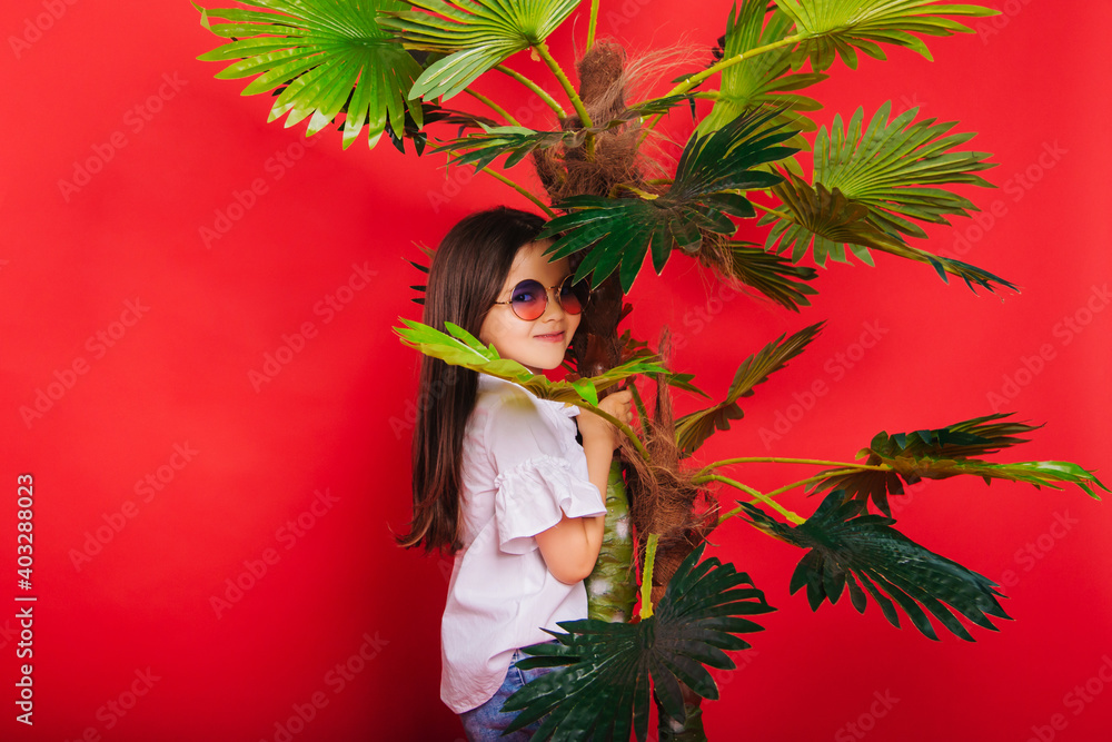 A little girl with round sunglasses stands next to a palm tree on a solid red background. A cheerful and energetic child. A little girl with round sunglasses stands next to a palm tree on a solid red background. A cheerful and energetic child.