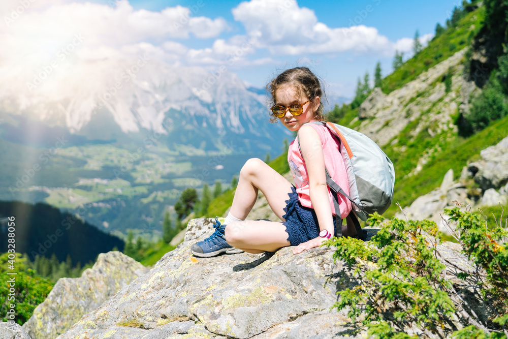 Naklejka premium Children hiking on beautiful summer day in alps mountains Austria, resting on rock and admire amazing view to mountain peaks. Active family vacation leisure with kids. Outdoor fun and healthy activity