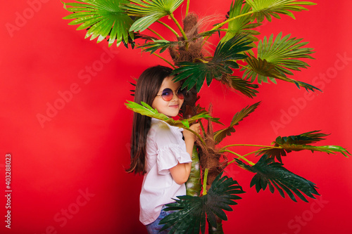 A little girl with round sunglasses stands next to a palm tree on a solid red background. A cheerful and energetic child. photo