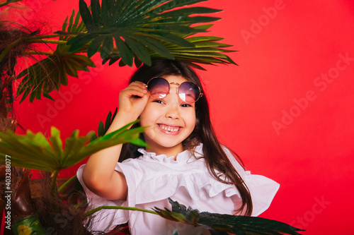 A little girl with round sunglasses stands next to a palm tree on a solid red background. A cheerful and energetic child. photo