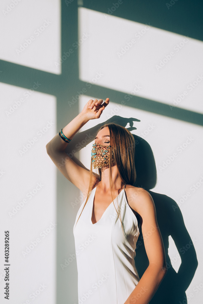 Naklejka premium Pandemic fashion. Female elegance. New normal lifestyle. Beautiful sensual woman in classic white dress designer golden chain turquoise beads face mask posing on sunlight.
