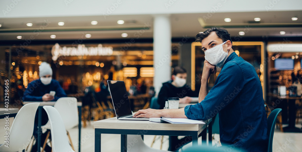 man in a protective mask works on a laptop sitting at a table in a cafe. man in a protective mask works on a laptop sitting at a table in a cafe.