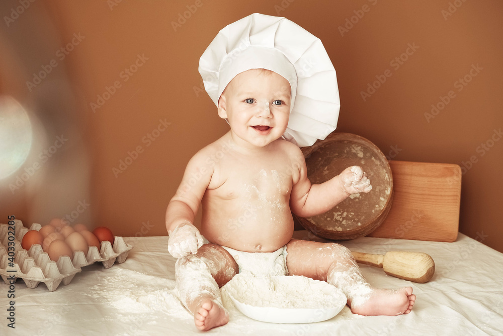 Little boy baker in a chef's hat sitting on the table playing with flour on a brown background with a wooden rolling pin, a round rustic sieve and eggs. Copy, empty space for text Little boy baker in a chef's hat sitting on the table playing with flour on a brown background with a wooden rolling pin, a round rustic sieve and eggs. Copy, empty space for text