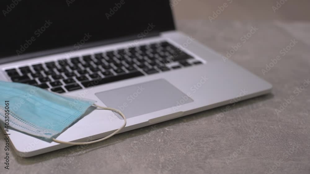 Computer laptop with medical face mask, notepad and pen on top of it together with a cup of coffee on dining table. Home setting. Work at home concept. Computer laptop with medical face mask, notepad and pen on top of it together with a cup of coffee on dining table. Home setting. Work at home concept.