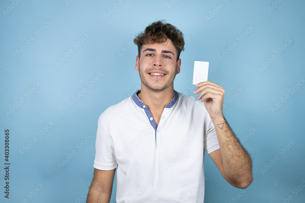 Young handsome man wearing a white t-shirt over blue background smiling and holding white card Young handsome man wearing a white t-shirt over blue background smiling and holding white card