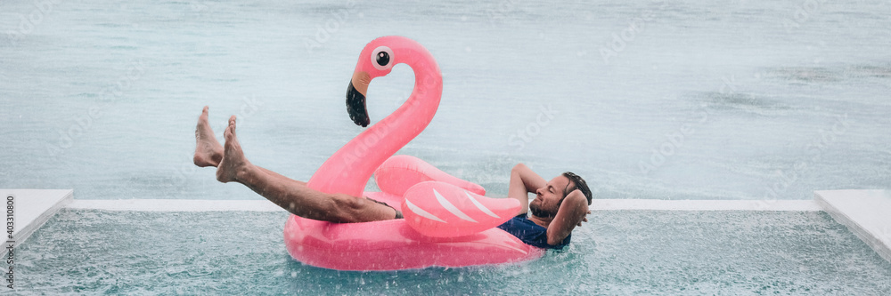 Happy man relaxing in swimming pool flamingo float despite bad rain weather. Travel summer vacation banner - obrazy, fototapety, plakaty 