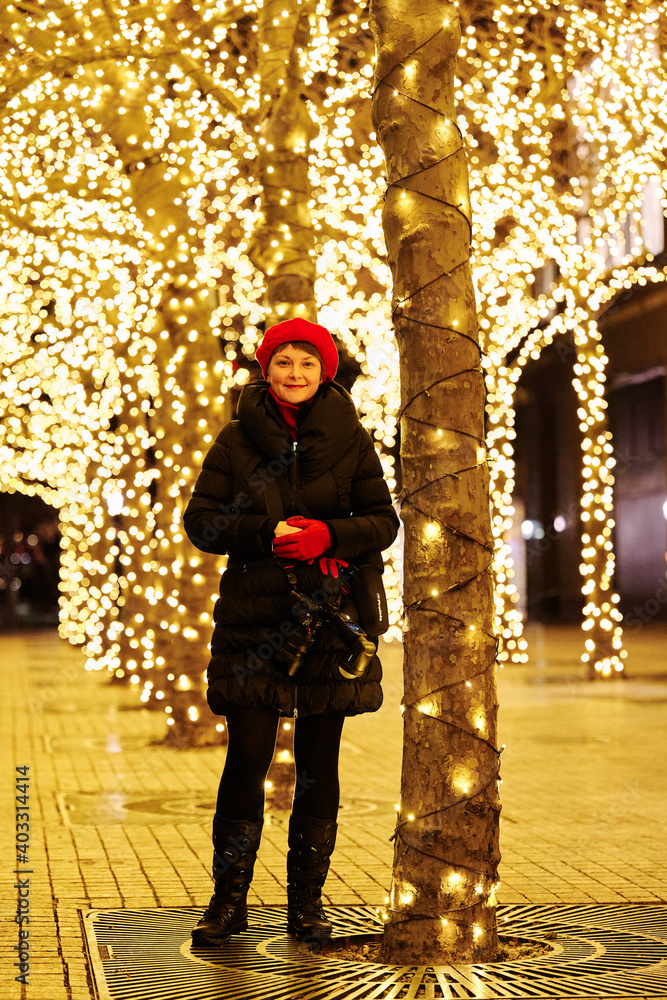 A woman is posing for a full-length portrait. It’s late at night and id dark outside. The Christmas Lights are in abundance in the background.