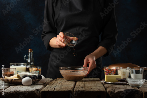 Pastry chef pours spaces into glass bowl for preparing dough on wooden table with variety of ingredients on dark blue background. Backstage of preparing sweet waffles. Frozen motion. Cooking process.