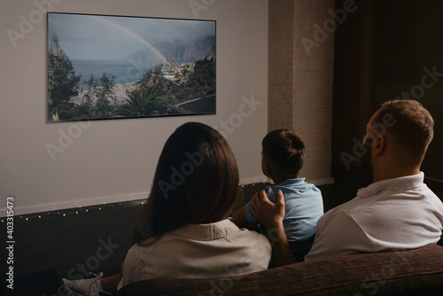 A photo from behind of a father, a son, and a young mother which are watching a movie on a widescreen television set on the sofa. The family is enjoying a video together in the evening at home.