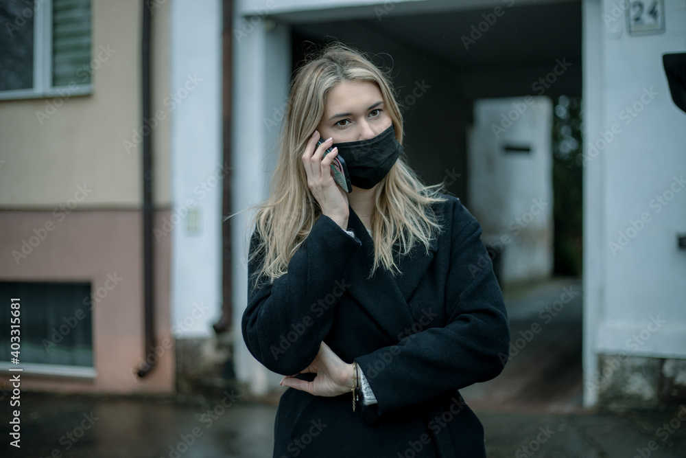 Fototapeta premium blonde business lady in black coat and mask on her face using smartphone against backdrop of arch of building. Lifestyle photo