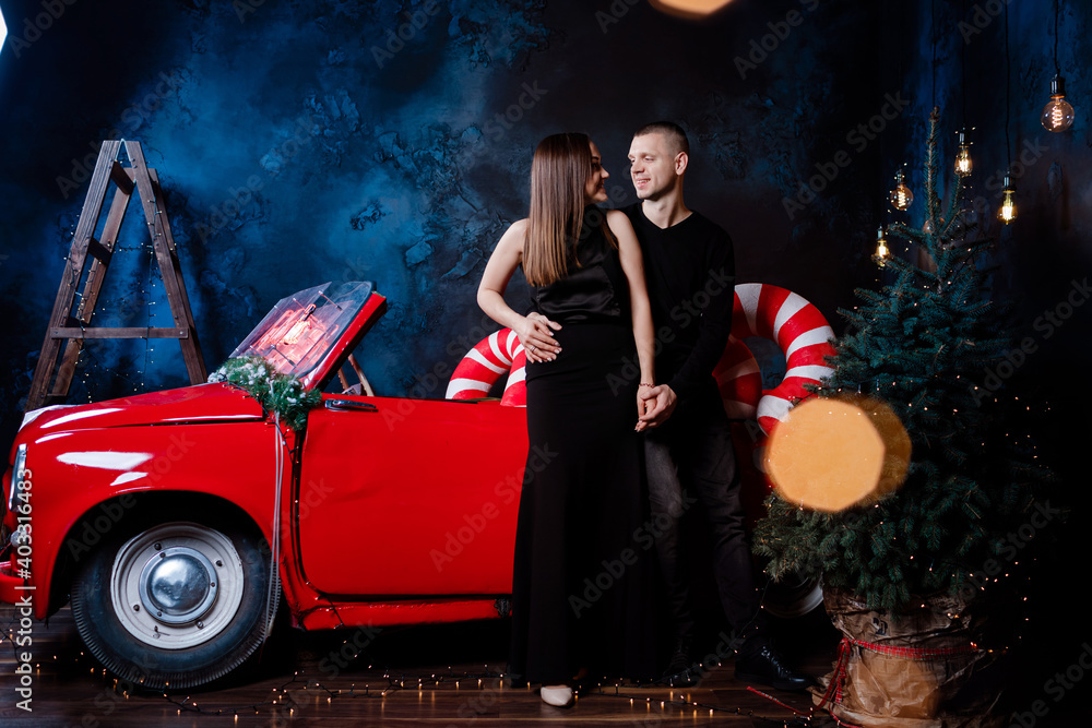 Young happy couple man and woman in love in Christmas are hugging near a red retro car with New Year's gifts. Kiss, girl, happiness, quarantine Christmas celebration, holiday.