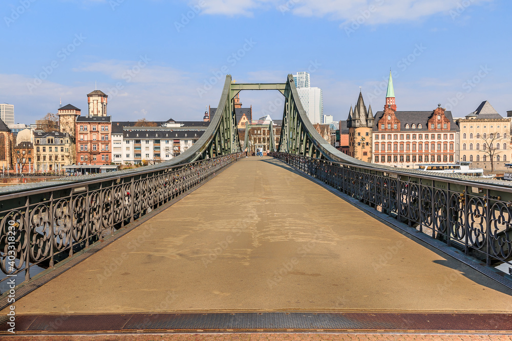 Obraz premium Historic footbridge over the Main in Frankfurt. Houses of the old town in the background with sunshine and a few clouds. Bridge with iron terrain