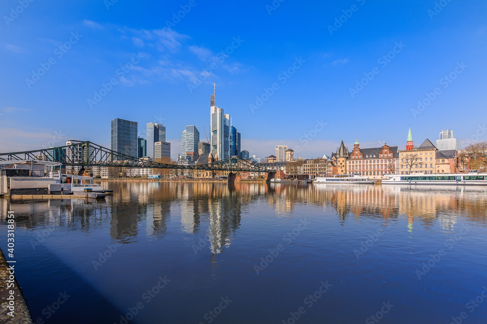 Financial district of Frankfurt in sunshine. Skyline commercial buildings and bridge over the river Main with reflections. Ships at the moorings and historical buildings under blue sky