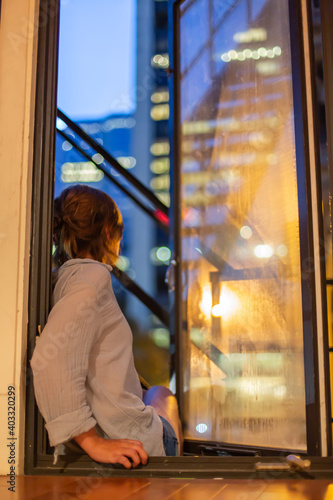 Aesthetic shot featuring a young woman sitting on a window ledge, taken from behind, as she gazes out on city lights of old Montreal port.