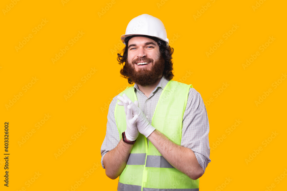 A portrait of an young bearded architect smiling is looking at the camera wearing a helmet some gloves and a phosphorescent vest .
